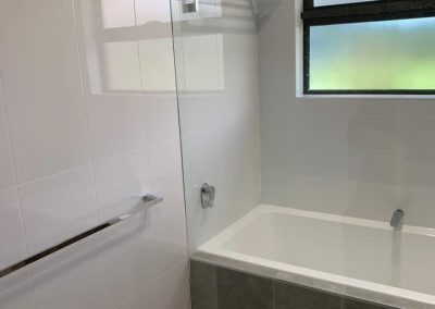 Built-in bath with a dark tiled front behind a frameless glass shower screen