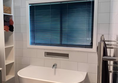 Freestanding bath under a window with blue blinds, beside a heated towel rail