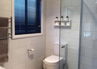 Toilet and frameless shower screen in a white-tiled bathroom with a window