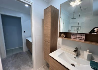 Tall timber storage cabinet beside a white vanity and mirror cabinet with timber shelf