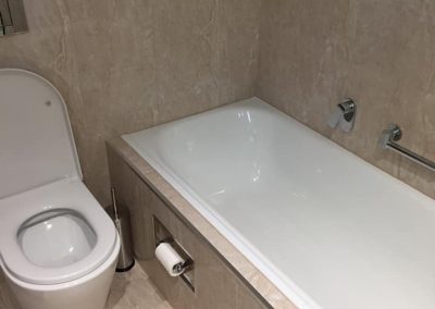 Built-in bath with a grab rail beside a toilet on beige marble-look tiles