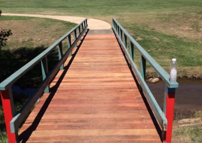 Timber footbridge with green and red railings over a creek on a grassed reserve