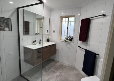 Black-framed shower screen and timber vanity in a white-tiled bathroom, wider view