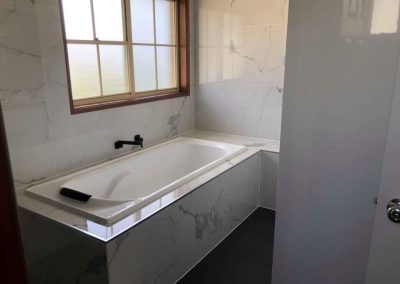 Built-in bath with marble-look tiled hob and black tapware under a window