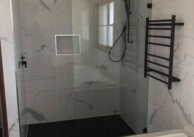 Walk-in shower with marble-look tiles, black rain shower and black towel ladder beside a bath