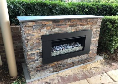 Outdoor fireplace with stacked stone cladding in front of a hedge