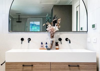Wide white basin with two sets of black wall taps and an arched black-framed mirror