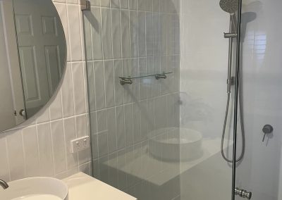 Frameless glass shower beside a white vanity with round vessel basin and round mirror