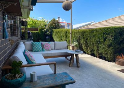 Outdoor lounge with cushions on a tiled patio beside a tall hedge