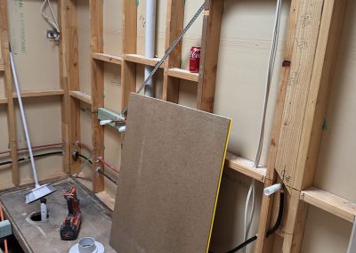 Gutted bathroom with exposed framing, pipework and a floor sheet propped against the wall