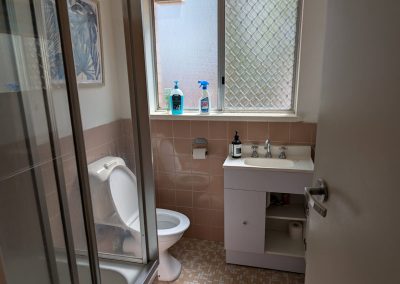 Dated bathroom with pink wall tiles, white vanity, toilet and glass shower screen